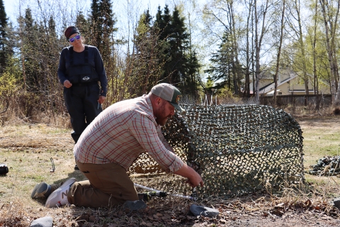 Two biologist building a camouflage canopy for the wooden raft with a tree line and houses in the background 