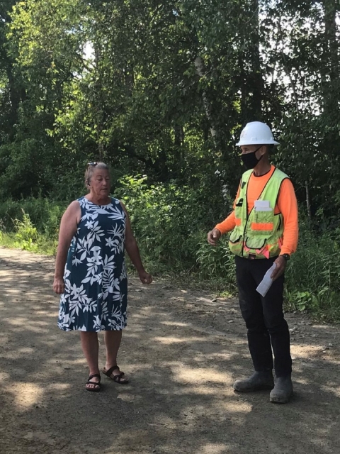 woman in blue dress talks to a person in a hard hat and reflective  vest on a dirt path in the woods on a sunny green day 