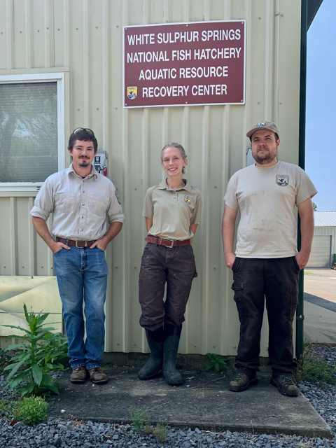 three people stand next to a building with a sign that reads White Sulphur Springs National Fish Hatchery Aquatic Resource Recovery Center