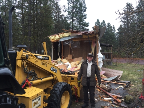 A man stands next to a bulldozer with a partially torn down building in the background. 