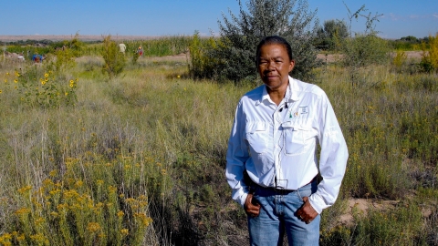 A woman with a white shirt and a lapel mic faces the camera with a desert wetland in the background