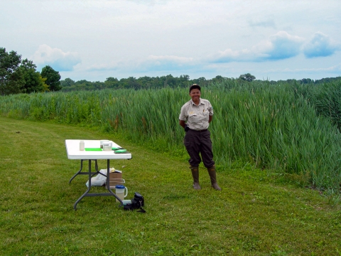 A biologist stands next to a folding table on green grass with tall wetland grass in the background.