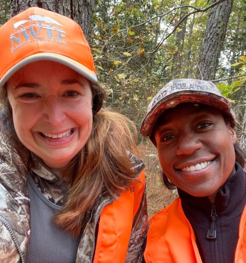 Two women wearing orange and camo, smile together in the woods