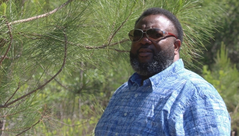A man in a blue button-down shirt with a dark bear and sunglasses standing in a pone forest setting