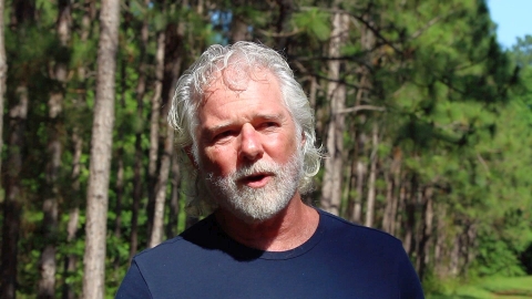 A man with long gray hair and a beard and wearing blue T-shirt standing front of a stand of trees