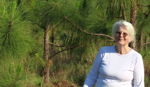 A woman in a white shirt standing in a pine forest setting 