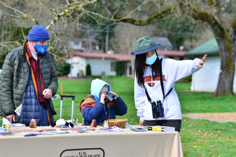 A Winter Wildlife volunteer staffs a USFWS table and shows a young child and their parent how to use binoculars.