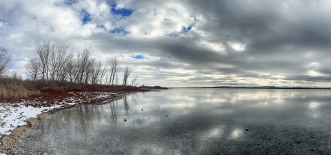 Clouds reflect on the surface of a lake, the shores are full of snow and trees without leaves.