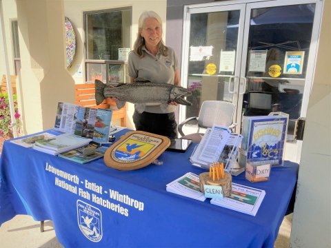 A fish hatchery volunteer holding a stuffed animal salmon stands behind a visitor information station in front of Leavenworth National Fish Hatchery