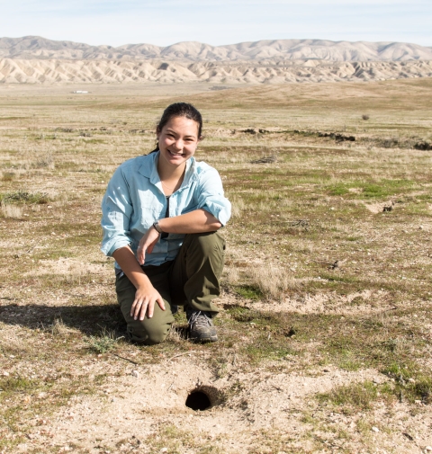 A woman kneels next to a small hole in the ground