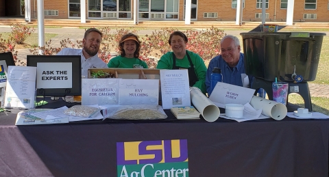 Two smiling women and two smiling men sit at an outdoor display with a wormbin