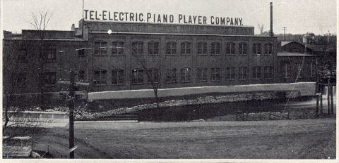 A black-and-white photo of a brick factory building with large capital letters spelling Tel-Electric Piano Player Company on top and a river flowing in the foreground