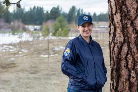 An adult woman wearing a blue ballcap and windbreaker stands next to a ponderosa pine.