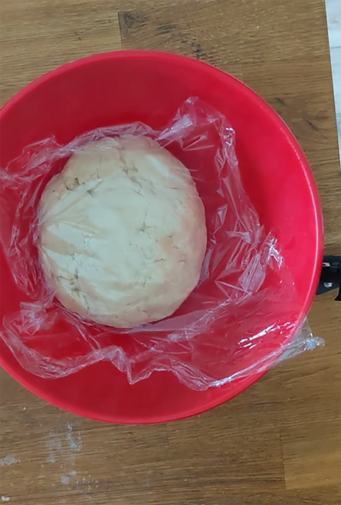 A ball of dough sitting in a red bowl wrapped in a piece of plastic wrap