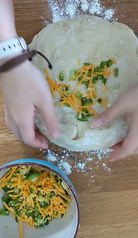 A pair of hands kneading a jalapeno and cheddar mixture into a ball of bread dough