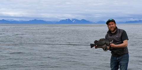 a man holding a black fish by the ocean with mountains in the background