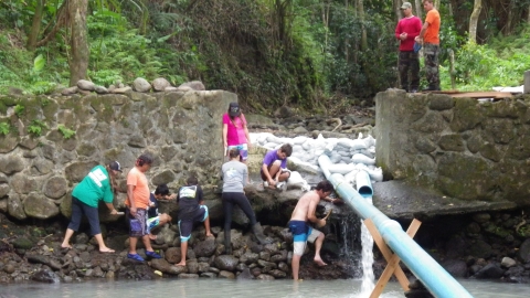 Several students in various colored clothes assist with fishway construction. A large tube stems up from a river to a smaller stream that is being directed with stones.