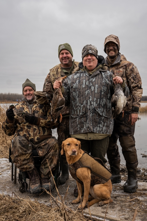 four adult hunters with wetland in background pose in camouflage hunting gear with dog 