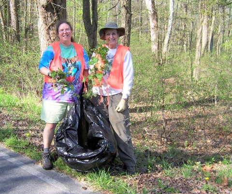 Two female volunteers standing in woods holding pulled garlic mustard plants and a large bag