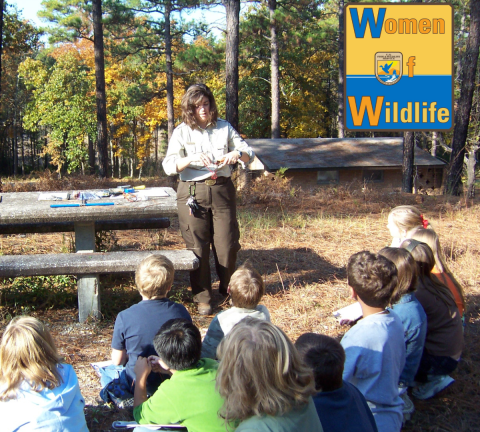 Allyne Askins stands in front of a group of students and teaches them about forestry. The Women of Wildlife logo is in the upper right corner.