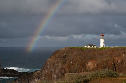 A rainbow arcs over the ocean and a lighthouse sitting on a rocky point. 