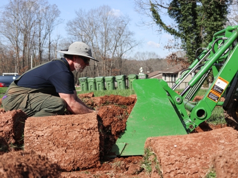 Person crouching in front of a tractor, lifting rolls of sod into the tracot bucket