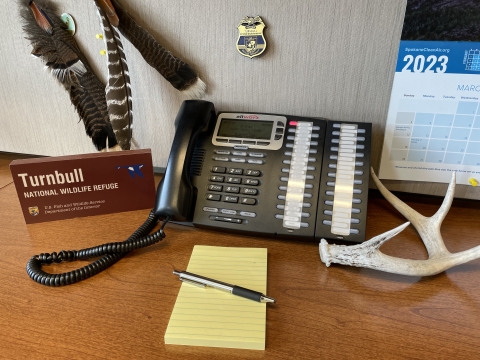 Photo of a telephone on a desk, with various items such as a pen, notepad, and calendar, surrounding it.