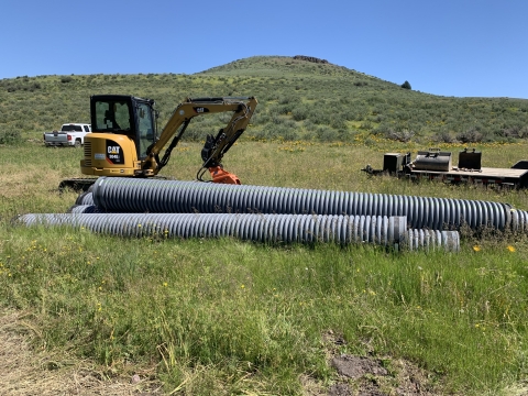 4 large gray pipes are seen in the grass in front of a backhoe machine.