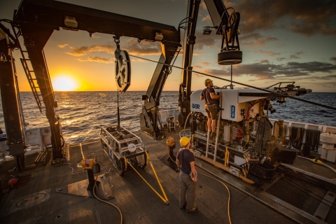 Crew work on a submersible on the deck of a large ship with the sun on the horizon of the ocean. 