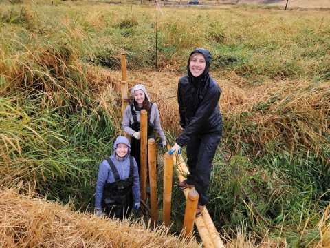 Three smiling young people outside building with wooden poles