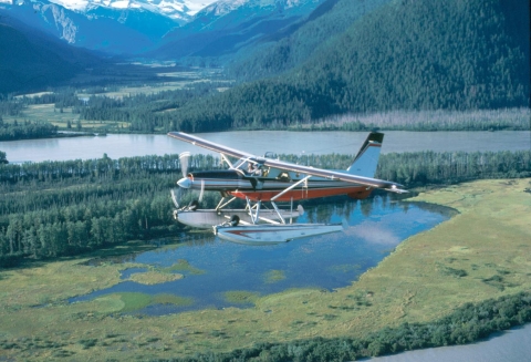 A small airplane flies above a lake with mountains in the background