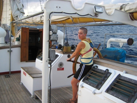 A woman with a shaved head steering a ship at sea looks over her shoulder at the camera.