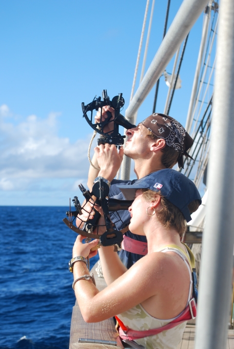 Two people look through navigational devices while standing along the rails of a ship on the ocean.