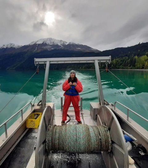 A woman wearing bright orange rain slicks and a blue life vest and hat stands on a fishing vessel in bright green water with a backdrop of snow-capped mountains.