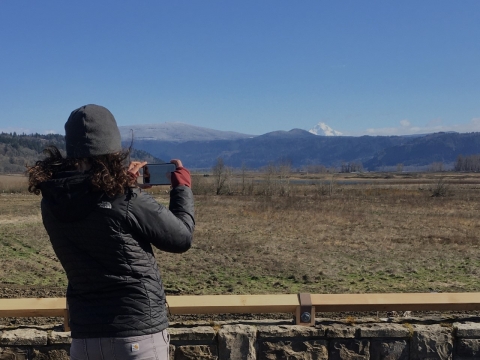 Visitor in a grey hat and pants and black coat uses cell phone to take a photo of the floodplain and Mt. Hood in the distance at the first overlook on the Mt. View Trail