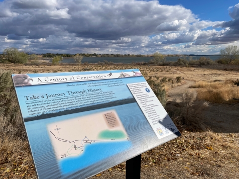 An interpretive sign about the history of the refuge with the lake and dramatic clouds in the background.