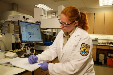 A woman wearing a white lab coat, gloves, and protective eyewear writes in a notebook while sitting in a laboratory.