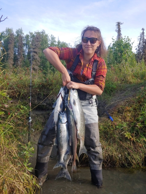 A woman wearing sunglasses, a plaid shirt, and waders holds up numerous fish while standing in a river.