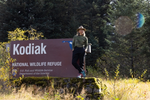 A woman stands by a brown sign with a blue goose logo that reads Kodiak National Wildlife Refuge.