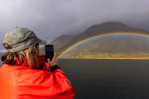 A woman uses her phone to take a photo of a rainbow across the mountains.