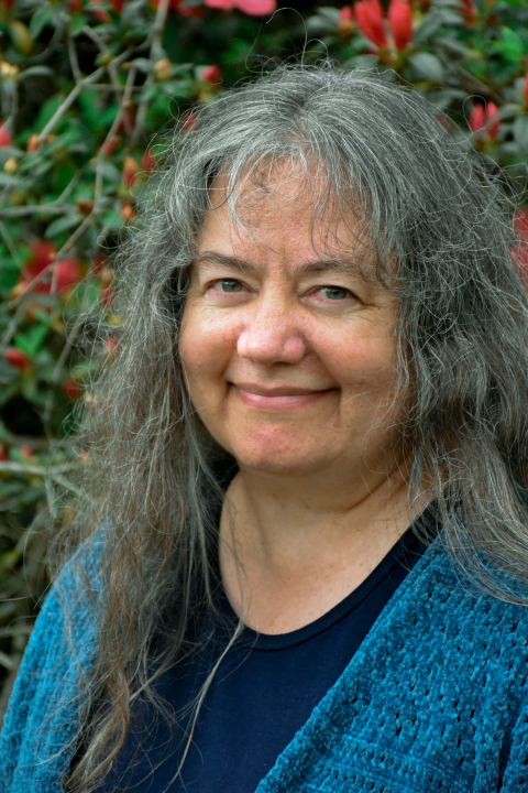 A woman with long, gray hair, blue top and blue sweater is smiling in front of a shrub with green leaves and orange flowers