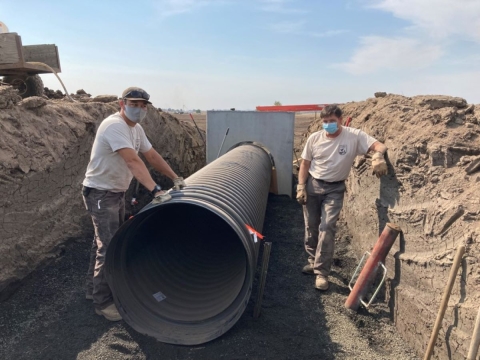 Two men in teeshirts and dust masks stand in a trench next to a large pipe. 