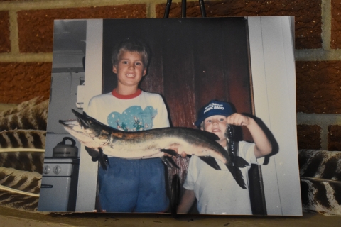 An old family photograph of two brothers; the biggest brother is holding a big fish and the little brother is holding a small fish.