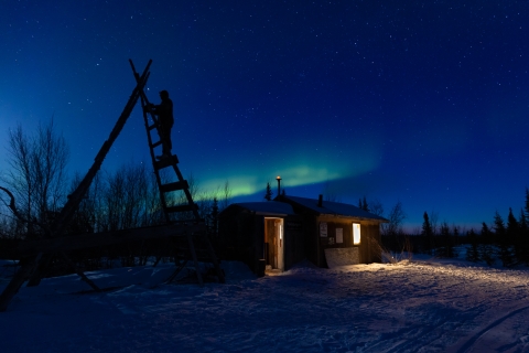 Green northern lights over a small cabin in the snow and the silhouette of a person on a lookout ladder.