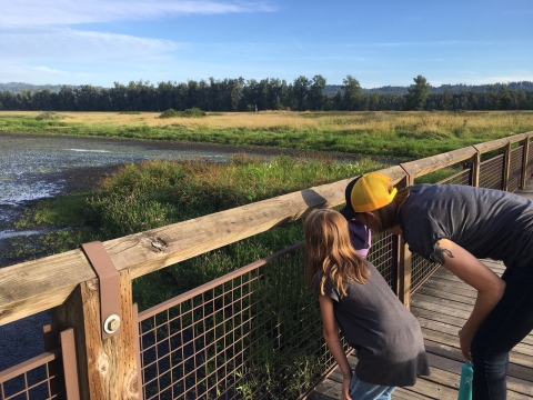 A child and a parent in a yellow hat peer between the railing of Redtail Bridge over Redtail Lake