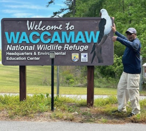 Stephen Meyers, a volunteer at Waccamaw National Wildlife Refuge, stands by the welcome sign at the entrance to the refuge and gestures excitedly with his hands.