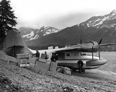 Three people offloading a Grumman N741 aircraft on a beach with mountains in the background. 