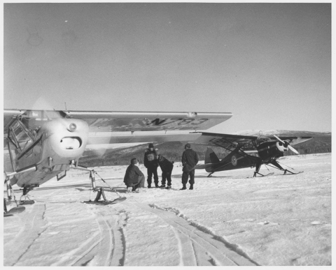 Four people stand on a snow covered ground in between two single engine propeller planes with skis equipped. 