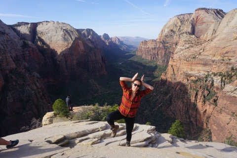 A woman on a cliff posing on one leg and hands up like antlers with a big canyon of red rocks and trees behind her.