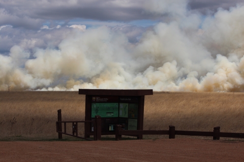 Smoke covers the sky behind a prairie and trail kiosk during a prescribed spring burn